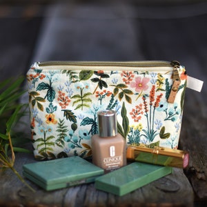 May include: A white floral print makeup bag with a gold zipper. The bag is open and contains a bottle of Clinique foundation, a gold lipstick, and two green makeup compacts.