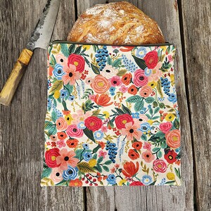 May include: A loaf of bread partially inside a floral bread bag. The bag features a colorful pattern of pink, red, blue, and yellow flowers with green leaves. A knife with a wooden handle rests on the wooden surface.
