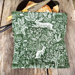 May include: A green and white fabric bread bag with a forest animal print, including rabbits and foxes. The bag is partially covering a cast iron skillet with a loaf of bread and a knife. The background is a rustic wooden surface.