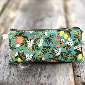 May include: A rectangular teal zippered pouch with a floral pattern featuring oranges, lemons, and white flowers. The pouch has a dark green zipper and a small cream-colored fabric pull tab. The background is a weathered wooden surface.