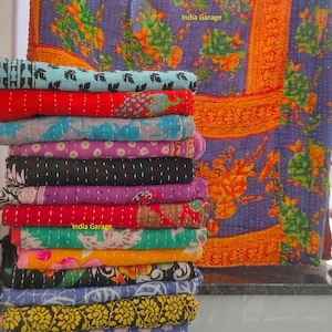 以下が含まれることがあります： 花柄のカラフルなカンタキルトの積み重ね。キルトは綿製で手縫いです。キルトは白い表面に積み重ねられ、背景には花柄のキルトが掛けられています。掛けられているキルトには「India Garage」の文字が見えます。