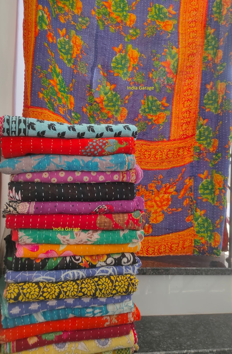 Peut inclure: Une pile de courtepointes kantha color&eacute;es avec des motifs floraux. Les courtepointes sont pli&eacute;es et empil&eacute;es les unes sur les autres. La courtepointe du dessus est un motif floral violet et orange avec une bordure dor&eacute;e. Le texte "India Garage" est visible sur la courtepointe du dessus.