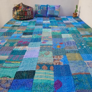 以下が含まれることがあります： 青と緑のパッチワークのキルトで、さまざまなパターンがあります。キルトはリサイクルされたサリーで作られており、手縫いです。キルトはベッドの上にあり、枕が2つと、テーブルの上に小さなサボテンがあります。