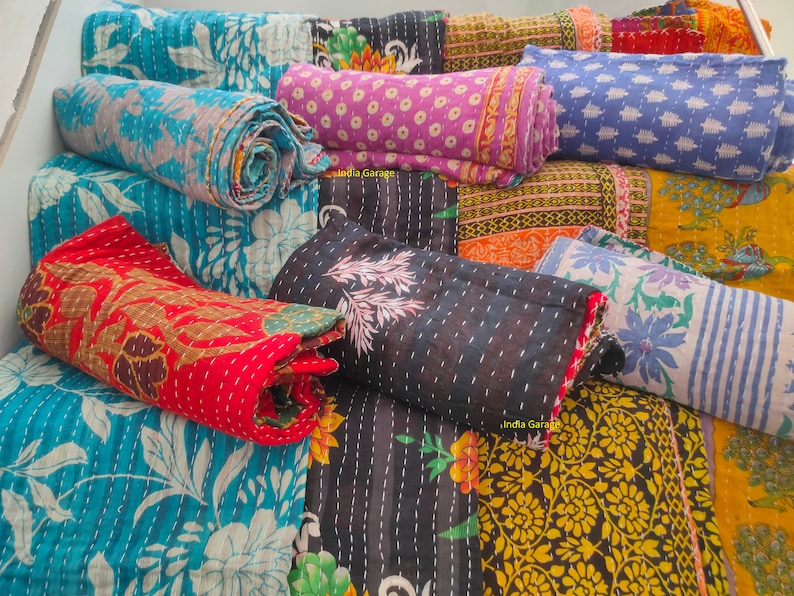 Peut inclure: Une collection de courtepointes kantha color&eacute;es, cousues &agrave; la main &agrave; partir de saris recycl&eacute;s, pr&eacute;sentant des motifs floraux et g&eacute;om&eacute;triques. Les courtepointes sont roul&eacute;es et dispos&eacute;es sur un escalier, mettant en valeur leurs d&eacute;tails complexes et leurs teintes vibrantes.