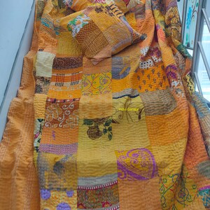 Op de afbeelding: Een patchworkquilt en twee bijpassende sierkussens in tinten geel, oranje en bruin. De quilt en kussens zijn gemaakt van verschillende stofresten die aan elkaar zijn genaaid. De quilt is over een oppervlak gedrapeerd.