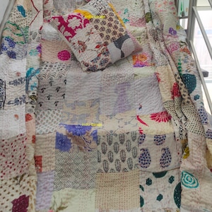 May include: A white kantha quilt with a patchwork design featuring various floral and geometric patterns in shades of blue, pink, red, and green. The quilt is made from recycled saris and is hand-stitched.