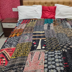May include: A black and white kantha quilt with a patchwork design. The quilt is made of various fabrics with different patterns and colors, including red, green, and brown. The quilt is laid out on a bed with white pillows and a red pillow.
