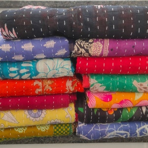 Peut inclure: Une pile de courtepointes kantha color&eacute;es avec des motifs floraux. Les courtepointes sont faites de saris recycl&eacute;s et sont cousues &agrave; la main. Les courtepointes sont dispos&eacute;es en pile verticale, la courtepointe du haut &eacute;tant noire et la courtepointe du bas &eacute;tant bleue. Le texte "India Garage" est visible sur deux des courtepointes.