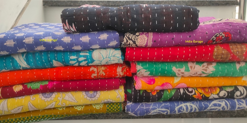 Peut inclure: Une pile de courtepointes kantha color&eacute;es avec des motifs floraux. Les courtepointes sont faites de saris recycl&eacute;s et sont cousues &agrave; la main. Les courtepointes sont dispos&eacute;es en pile verticale, la courtepointe du haut &eacute;tant noire et la courtepointe du bas &eacute;tant bleue. Le texte "India Garage" est visible sur deux des courtepointes.