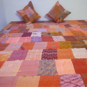 Könnte beinhalten: Eine Patchwork-Steppdecke und zwei passende quadratische Kissen. Die Steppdecke zeigt eine Vielzahl warmer Farben, darunter Orange, Rosa und Braun, die in einem geometrischen Muster angeordnet sind. Die Kissen greifen das Design der Steppdecke auf.