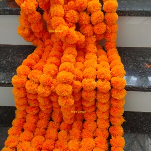 Puede incluir: Guirnaldas de flores de caléndula de color naranja brillante cayendo en cascada sobre una superficie oscura. Las guirnaldas están densamente repletas de flores esféricas vibrantes, creando una exhibición rica y texturizada. La imagen incluye el texto "India Garage".
