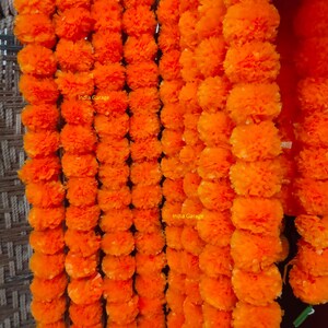 May include: Rows of vibrant orange marigold garlands. The garlands are densely packed with fluffy, round blooms, creating a full and textured appearance. The text "India Garage" is visible on the garlands.