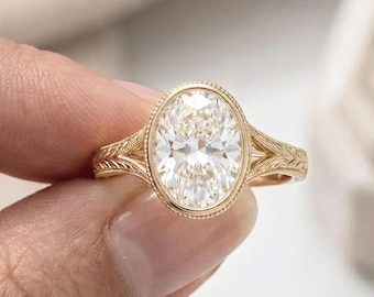 May include: A gold engagement ring with an oval-cut diamond. The ring features a detailed filigree design on the band and around the diamond setting. The diamond sparkles brightly, reflecting light. The ring is held up by a person's fingers.