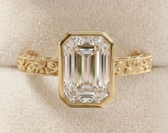 Pode incluir: Um anel de ouro com uma grande pedra preciosa transparente de corte esmeralda retangular engastada num bisel. A banda é feita de ouro amarelo e apresenta um padrão intrincado de volutas florais em relevo nas laterais do metal.