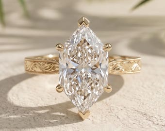 Op de afbeelding: Een gouden ring met een grote, heldere edelsteen in marquise-slijpvorm, vastgehouden door zes klauwen. De ringband is gemaakt van geelgoud en is voorzien van een ingewikkeld, gegraveerd krulpatroon over de gehele buitenzijde.