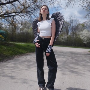 May include: A person wearing a white crop top, black jeans, and sneakers, posing outdoors. The person is wearing large, dark gray angel wings and striped arm warmers. The background includes trees and a paved path.