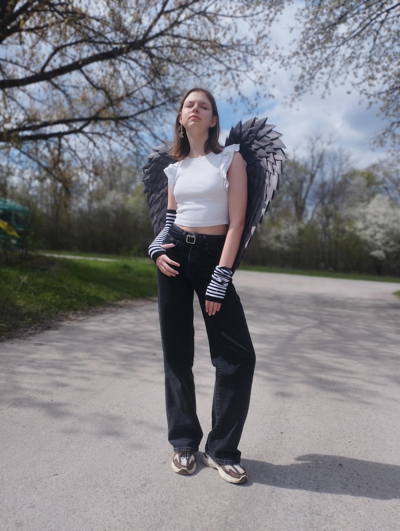 May include: A person wearing a white crop top, black jeans, and sneakers, posing outdoors. The person is wearing large, dark gray angel wings and striped arm warmers. The background includes trees and a paved path.
