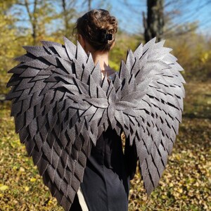 May include: Gray, layered wing costume with a textured appearance. The wings are designed to be worn on the back, with a central spine and individual feather-like elements. The costume is set against a backdrop of fall foliage.