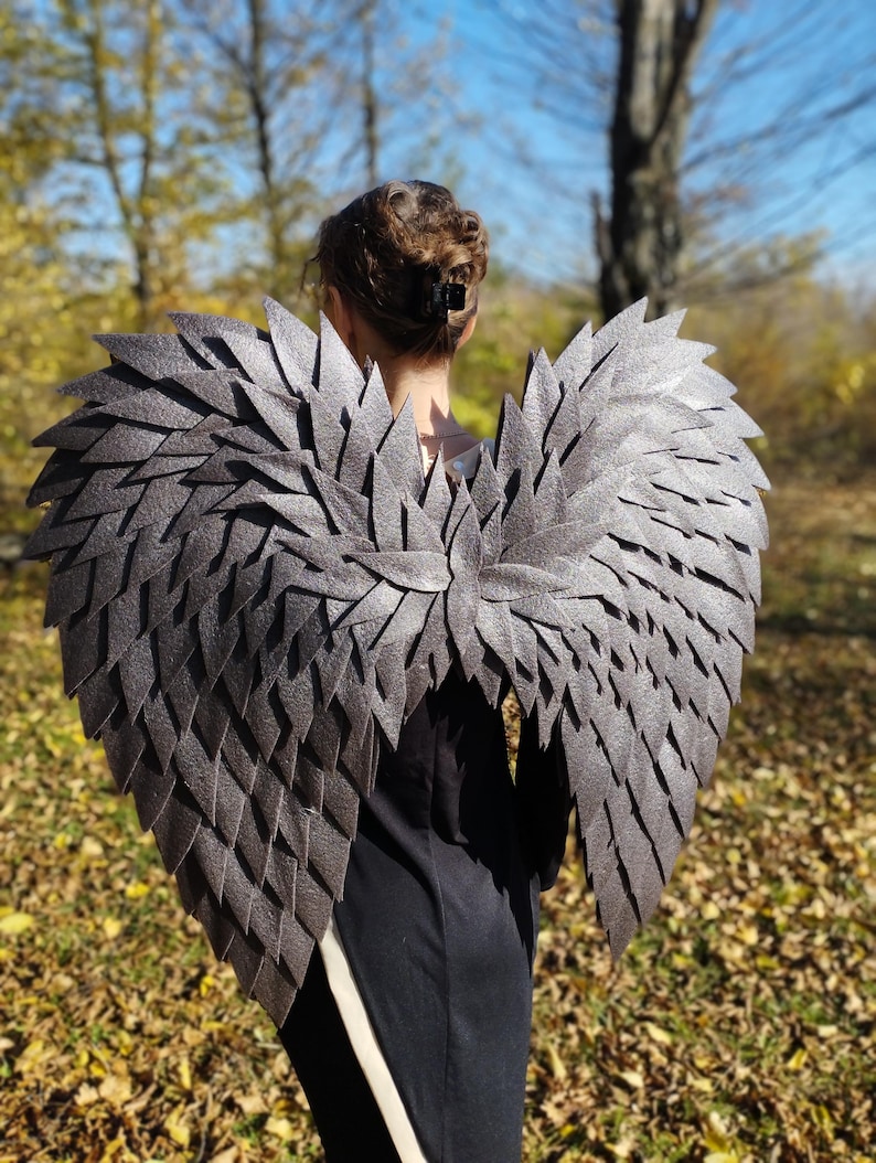 May include: Gray, layered wing costume with a textured appearance. The wings are designed to be worn on the back, with a central spine and individual feather-like elements. The costume is set against a backdrop of fall foliage.
