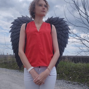 May include: A person wearing a red sleeveless top and white pants, with large black feathered wings attached to their back. The person is standing on a dirt path under a cloudy sky.