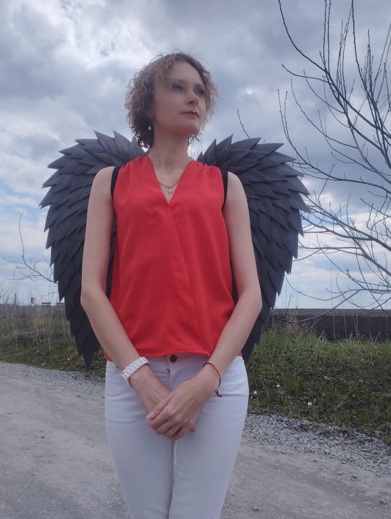 May include: A person wearing a red sleeveless top and white pants, with large black feathered wings attached to their back. The person is standing on a dirt path under a cloudy sky.