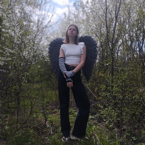 May include: A person wearing black angel wings, a white top, black pants, and striped arm warmers stands in a forest. The wings are large and dark, contrasting with the white blossoms in the background. The outfit suggests a costume or cosplay theme.