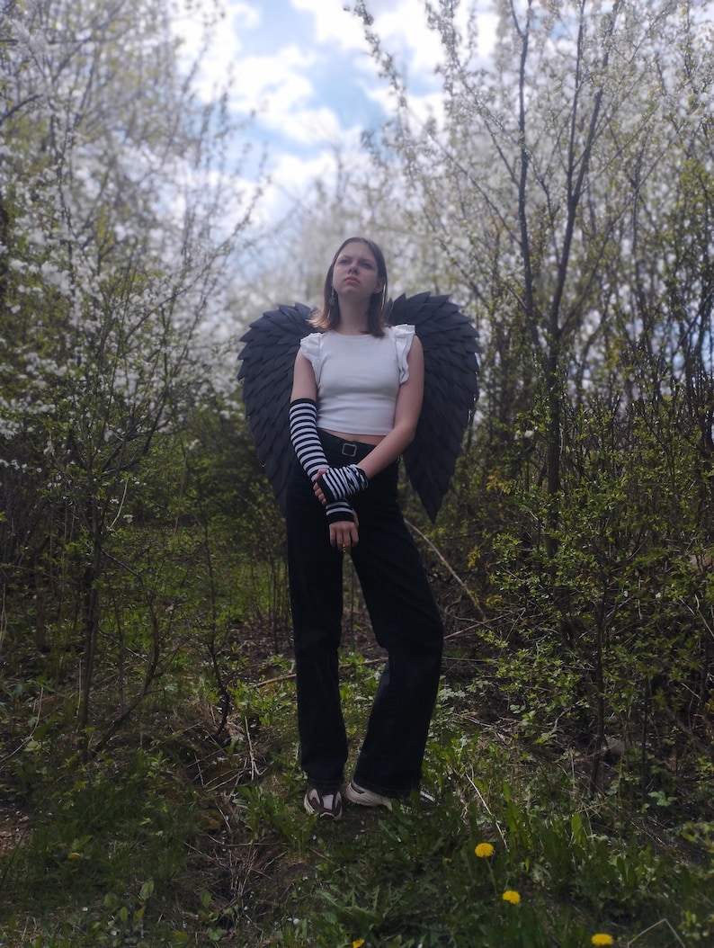 May include: A person wearing black angel wings, a white top, black pants, and striped arm warmers stands in a forest. The wings are large and dark, contrasting with the white blossoms in the background. The outfit suggests a costume or cosplay theme.