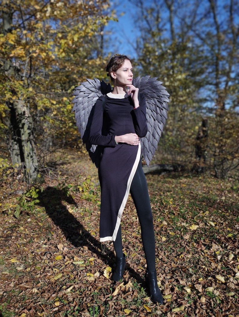 May include: A person wearing a black dress with a white accent and black tights, posing outdoors. The person is wearing large, gray, feathered angel wings. The background features trees with yellow and green leaves, and a blue sky.