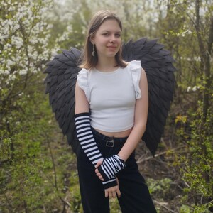 May include: A person wearing a white crop top, black pants, and black and white striped arm warmers. Large, dark gray angel wings are attached to the back. The background features green foliage and white flowering trees.