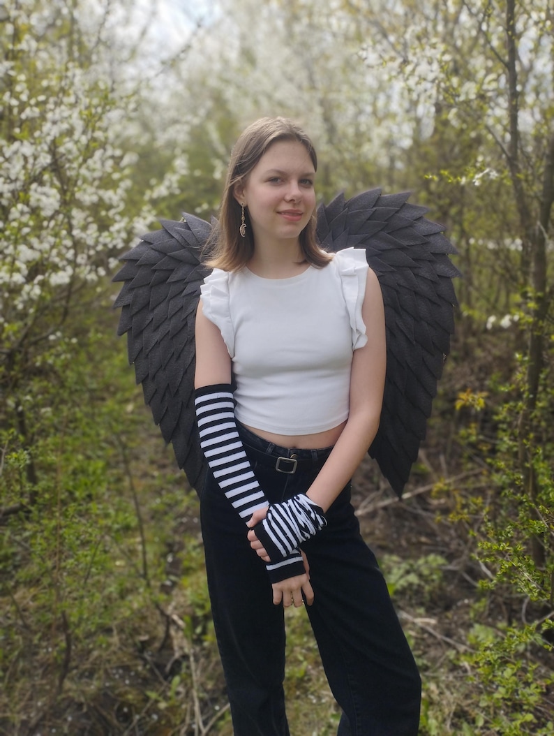 May include: A person wearing a white crop top, black pants, and black and white striped arm warmers. Large, dark gray angel wings are attached to the back. The background features green foliage and white flowering trees.