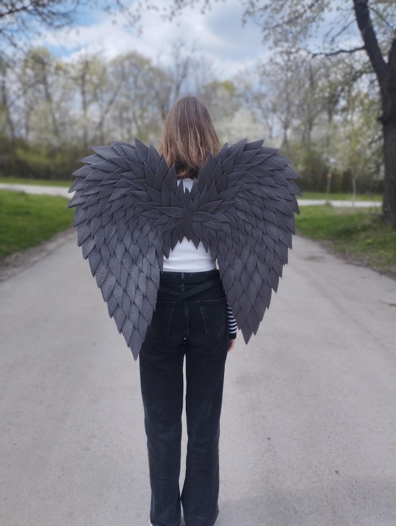 May include: Dark gray, layered feather-like wings. The wings are worn on the back, with a person facing away from the camera. The wings are made of a textured material and are designed to resemble a bird's wings. The person is wearing black jeans and a white shirt.