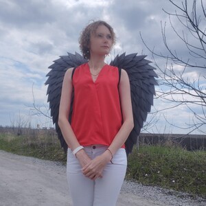 May include: A person wearing a red top, white pants, and dark gray angel wings stands on a gravel road. The wings are large and detailed, with a layered feather design. The sky is overcast.