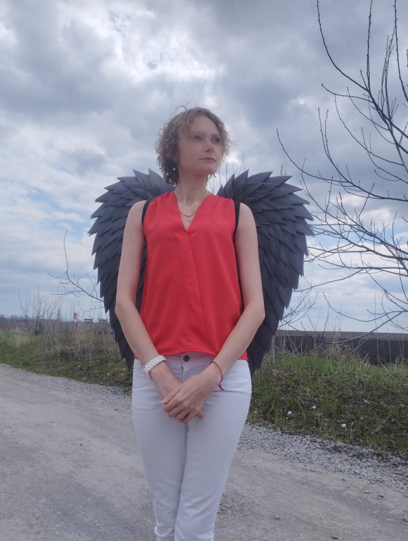 May include: A person wearing a red top, white pants, and dark gray angel wings stands on a gravel road. The wings are large and detailed, with a layered feather design. The sky is overcast.