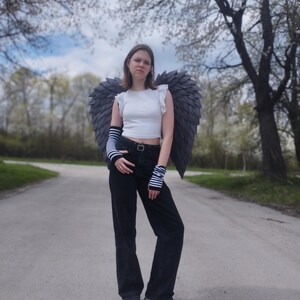 May include: A person wearing a white crop top, black jeans, and gray angel wings stands on a paved path. The wings are large and detailed, with a layered feather design. The person is also wearing striped arm warmers and sneakers.