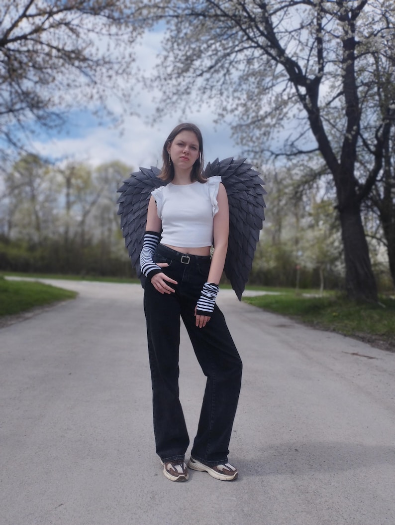 May include: A person wearing a white crop top, black jeans, and gray angel wings stands on a paved path. The wings are large and detailed, with a layered feather design. The person is also wearing striped arm warmers and sneakers.