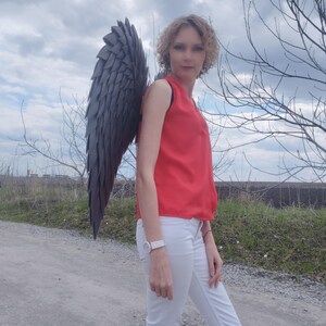 May include: A person wearing a red sleeveless top and white pants stands outdoors, with large, dark gray feathered wings attached to their back. The wings are the focal point, with a layered design.