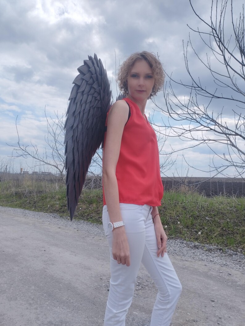 May include: A person wearing a red sleeveless top and white pants stands outdoors, with large, dark gray feathered wings attached to their back. The wings are the focal point, with a layered design.