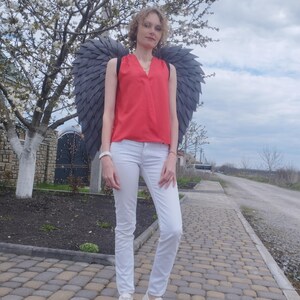May include: A person wearing a red sleeveless top, white pants, and white sneakers, with large gray angel wings. The person is standing on a brick-paved walkway in front of a flowering tree.