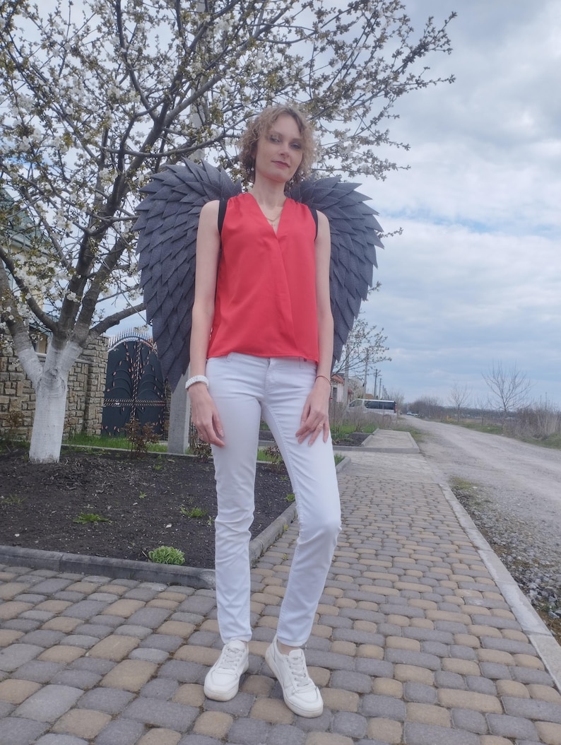 May include: A person wearing a red sleeveless top, white pants, and white sneakers, with large gray angel wings. The person is standing on a brick-paved walkway in front of a flowering tree.