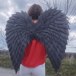 May include: A person wearing large, gray felt angel wings. The wings are made of layered, pointed felt pieces, and are attached to the person's back. The person is wearing a red top and white pants.