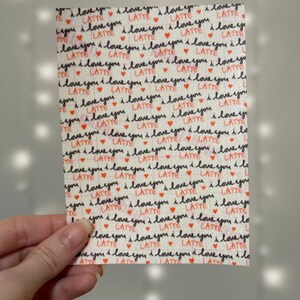 Puede incluir: Una tela blanca con un patr&oacute;n repetido de las palabras "Te amo" y "Latte" en escritura negra con peque&ntilde;os corazones rojos.