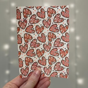 Puede incluir: Una hoja de tela blanca con un patr&oacute;n repetido de galletas con forma de coraz&oacute;n rojas y marrones. Los corazones tienen un fondo blanco y un contorno marr&oacute;n.