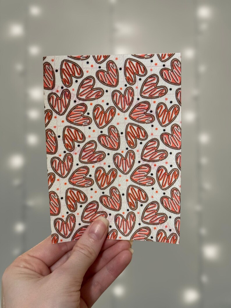 Puede incluir: Una hoja de tela blanca con un patr&oacute;n repetido de galletas con forma de coraz&oacute;n rojas y marrones. Los corazones tienen un fondo blanco y un contorno marr&oacute;n.