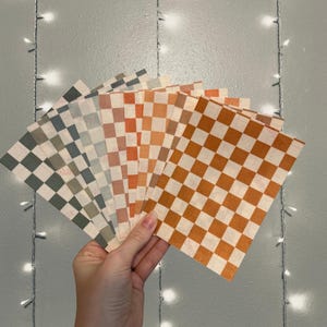 May include: A hand holds a fan of square napkins in various colors. Each napkin features a checkered pattern in shades of green, gray, orange, and brown. The background is a light gray wall with string lights.