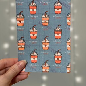 Puede incluir: Una tarjeta con un patr&oacute;n azul y blanco con un patr&oacute;n repetido de vasos de caf&eacute; helado naranja y blanco con un coraz&oacute;n en el lateral. El texto "Te amo" se repite en todo el patr&oacute;n.