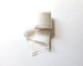 Op de afbeelding: Een wit zijden lint met rafelige randen, opgerold en klaar voor gebruik bij het knutselen of decoreren.