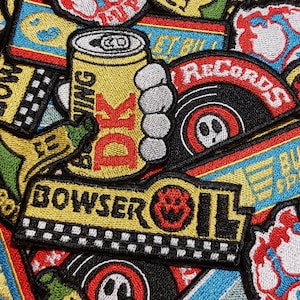 May include: A collection of embroidered patches featuring various designs including a yellow can labeled DK, a Bowser Oil logo with a checkered pattern, a banana, and circular records with red and black details, all arranged in a dense overlapping pattern.