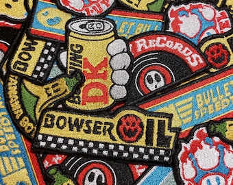 May include: A collection of embroidered patches featuring various designs including a yellow can labelled DK, a Bowser Oil logo with a checkered pattern, a banana, and circular records with red and black details, all arranged in a dense overlapping pattern.