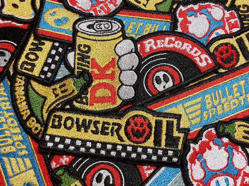 May include: A collection of embroidered patches featuring various designs including a yellow can labeled DK, a Bowser Oil logo with a checkered pattern, a banana, and circular records with red and black details, all arranged in a dense overlapping pattern.