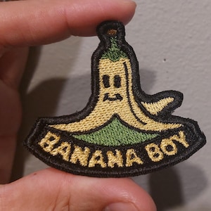 May include: An embroidered patch featuring a cartoon banana character with a yellow peel and green stem. The character has a simple face with two eyes and a mouth. Below the character, the text BANANA BOY is stitched in yellow on a black background.
